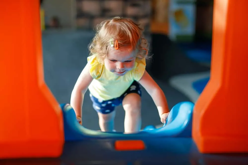 Happy blond little toddler girl having fun and sliding on indoor playground at daycare or nursery. Positive funny baby child smiling. Healthy girl climbing on slide