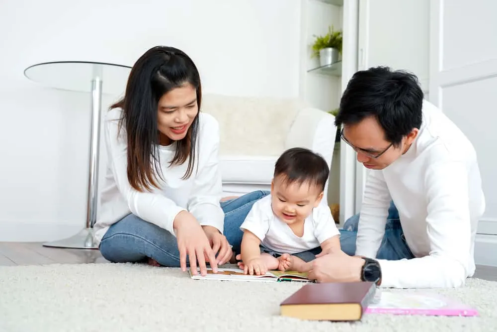 Happy modern Asian parents sitting on floor with cute infant boy and reading fairy tale while enjoying time together at home